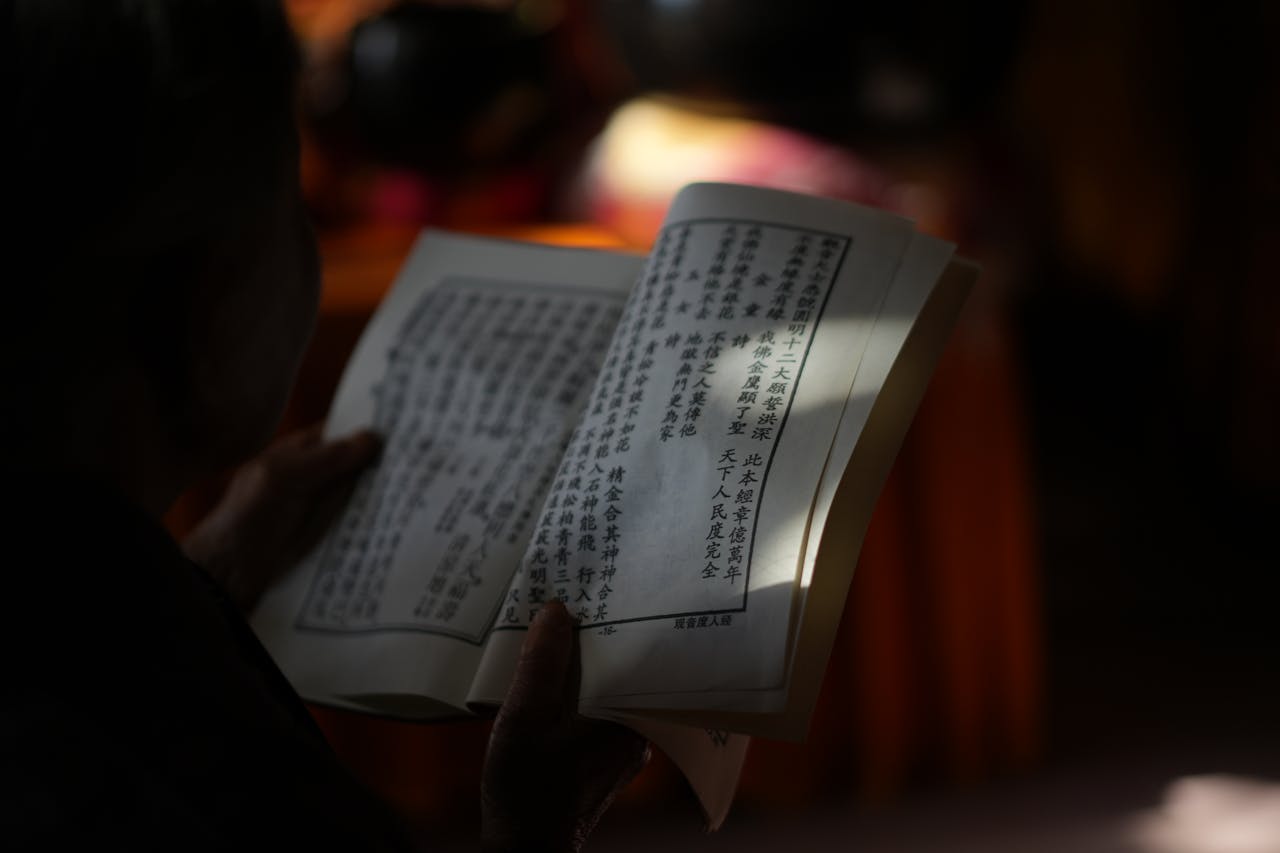 Kodu Close-up of hands holding a Chinese book, illuminated softly amidst shadows, emphasizing reading focus.